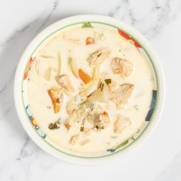 Creamy chicken soup with carrots, onions, and herbs in a decorative bowl on a marble surface.