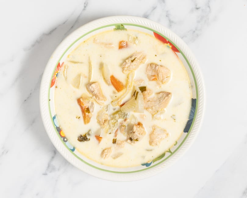 Creamy chicken soup with carrots, onions, and herbs in a decorative bowl on a marble surface.