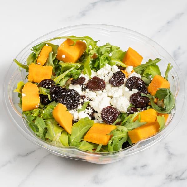 A clear plastic bowl filled with a salad of green leaves, orange butternut squash, white feta cheese, and dark dried cranberries.