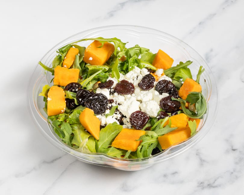 A clear plastic bowl filled with a salad of green leaves, orange butternut squash, white feta cheese, and dark dried cranberries.