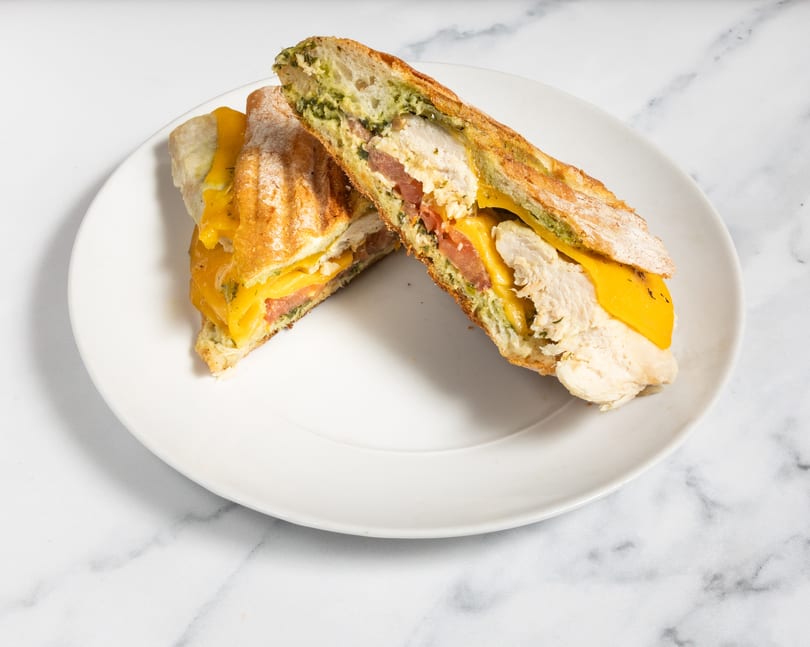 Two halves of a chicken panini with melted cheese, tomatoes, and pesto on a white plate.